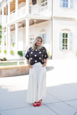 Woman posing outdoors in the Chocolate Polka Dot Knit Sweater, a playful yet chic short-sleeve top featuring large white polka dots, a classic crew neckline, and a relaxed fit. The knit sweater is paired with flowy cream wide-leg pants featuring a ruffled paperbag waist and front patch pockets. Styled for a vibrant, transitional aesthetic with bold red strappy platform sandals and layered statement jewelry, standing on a paved walkway in front of an elegant white building with classical columns and light bl