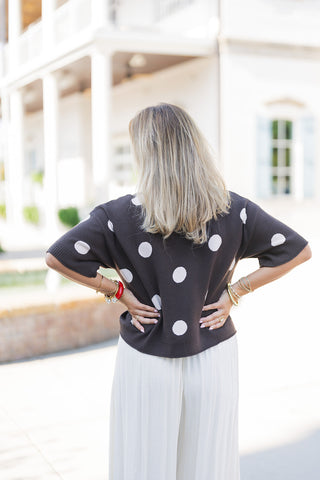 Back view of the Chocolate Polka Dot Knit Sweater showcasing the seamless continuation of the bold polka dot print across the shoulders and bodice. The slightly cropped, ribbed hem rests cleanly over the waistband of the cream wide-leg trousers. With her hands resting on her hips, the image highlights the relaxed, comfortable silhouette of the knit top and the elegant, fluid pleats of the trousers.