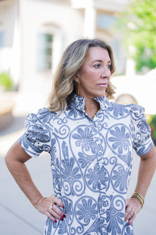 A portrait view of the Blue Botanical Scroll Midi Dress highlighting the textured detail of the ruffled high collar and the classic white button-down placket. The close-up emphasizes the rich blue printed fabric and the gathered puff sleeves adorned with delicate embroidered eyelet detailing. The look is accessorized with elegant layered gold necklaces, rings, and stacked bracelets.