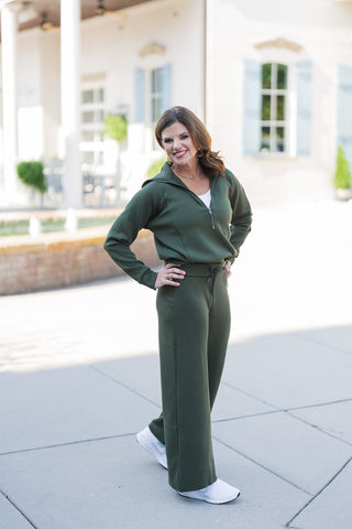 Woman confidently posing outdoors in the Mossy Wide Leg Pant, styled as part of a matching olive green loungewear set. The pants feature a comfortable high-waisted fit with an adjustable drawstring, paired perfectly with a coordinating half-zip long-sleeve pullover layered over a white tee. The relaxed, wide-leg silhouette falls effortlessly over white athletic sneakers, creating a chic, sporty aesthetic in front of a classic white building with light blue shutters.