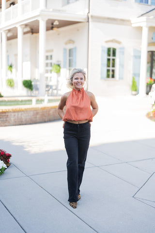 Woman posing outdoors in the Rust Satin Halter Top, a silky burnt orange blouse featuring a high, ruched neckline and a softly draped bubble hem. She is styled for a chic, transitional look, pairing the slightly cropped top with black straight-leg jeans that have subtle rhinestone detailing down the side seams, and pointed-toe leopard print shoes. She stands smiling with her hands on her hips in front of an elegant white building with classical columns and light blue shutters.