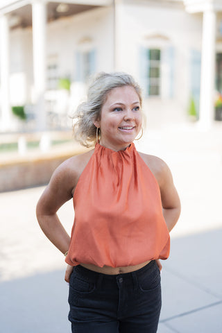 A portrait view of the Rust Satin Halter Top highlighting the lustrous, fluid texture of the fabric and the architectural detail of the gathered high neckline. The top drapes effortlessly into an elasticized banded hem that rests just above the waistline of the black denim. Accessorized with large gold hoop earrings and a relaxed curly updo, creating a warm, vibrant aesthetic.