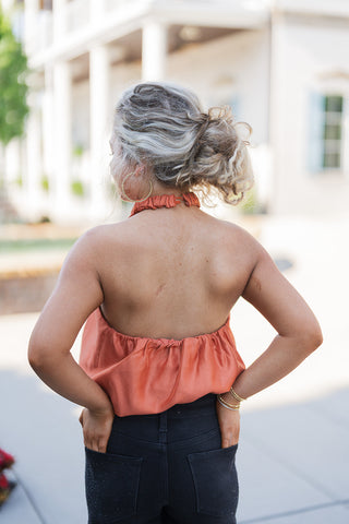 Back view of the Rust Satin Halter Top showcasing the daring open-back silhouette. The image highlights the ruched neckband with a clasp closure at the nape of the neck, leaving the shoulders and mid-back completely bare. The fabric gathers cleanly at the lower back into the bubble hem, seamlessly meeting the waistband of the black jeans for a sophisticated, night-out ready fit.