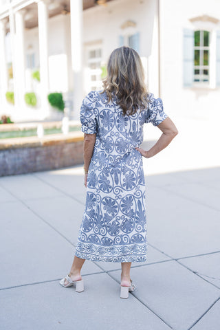 Back view of the Blue Botanical Scroll Midi Dress showcasing the relaxed, breathable silhouette and the continuous flow of the intricate blue botanical print. The midi-length design highlights the structured shoulders of the puff sleeves and features a distinct contrasting border pattern along the bottom hem, finished with subtle eyelet cutouts for an airy, elevated look.