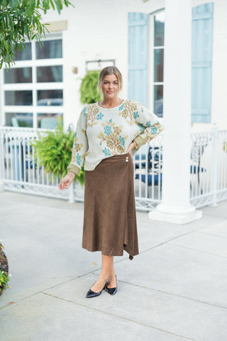 Woman posing outdoors wearing an oversized cream crewneck knit sweater decorated with a teal, olive green, and taupe botanical floral tree pattern. The sweater features dropped shoulders and ribbed cuffs. She is styled with a rich brown faux-suede wrap midi skirt featuring decorative side gold buttons, paired with black pointed-toe slingback flats and delicate gold jewelry.
