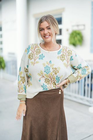 A waist-up portrait emphasizing the soft texture and knit construction of the cream crewneck sweater with its muted blue, green, and tan floral branch motif. Details show the ribbed neckline, turned-up cuffs, gold button accents on the brown suede wrap skirt waist, a dainty gold pendant necklace, and stacked gold bracelets.