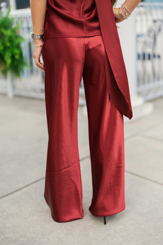 Back view detailing the rear fit of the burgundy satin trousers, highlighting a smooth, darted waist design and wide-leg hem that cascades gracefully over dark heels. Showcases the drape of a matching neck-tie sash from the halter top hanging along the right side, styled with delicate beaded wrist bracelets and a watch.