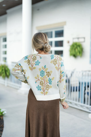 Back view showcasing the continuous botanical floral knit design across the reverse side of the relaxed cream sweater. The sweater features a clean ribbed hem paired with the smooth back silhouette of the brown suede midi skirt. The model wears her hair in a low, textured bun.