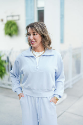 Woman posing outdoors wearing a light blue half-zip sweatshirt featuring a wide fold-over collar, structured bib-stitching across the chest, long sleeves with ribbed cuffs, and a wide ribbed waistband hem. The athleisure pullover is layered over a white tee and paired with matching light blue drawstring wide-leg lounge pants, styled with a gold chain necklace, drop earrings, and stacked gold bangles against a white storefront backdrop.
