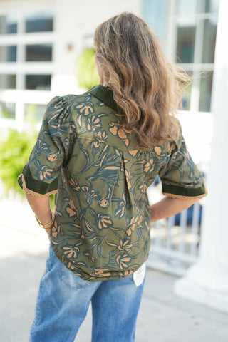 Back view of the olive green floral print blouse showing the back yoke with a central box pleat for an easy, comfortable drape. Highlights the dark green corduroy-trimmed sleeve cuffs with tan scalloped lace borders and subtle puff shoulder detailing, styled with light-blue jeans.
