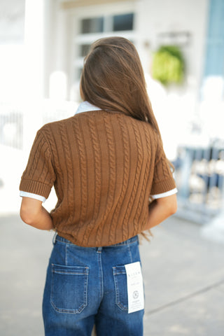 Back view of the short-sleeve cable-knit sweater showing the continuous vertical cable design across the back, ribbed waist hem, and ribbed sleeve cuffs with subtle white layering visible at the collar and sleeves. Styled with dark blue jeans with back patch pockets.