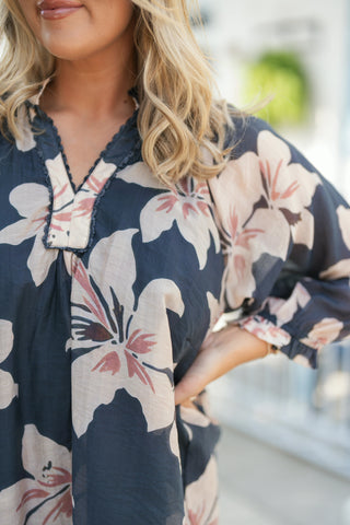 A portrait view of the navy floral blouse highlighting the intricate dark picot trim detailing along the split V-neck placket. Showcases the lightweight, semi-sheer woven fabric texture, the soft blush-pink floral motifs, and the gentle gathers along the neckline and ruffle cuffs.