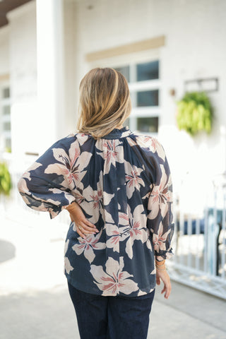 Back view of the navy and blush floral blouse showcasing the gathered, ruffled stand collar and gathered back yoke that creates a fluid, comfortable drape. Highlights the three-quarter length sleeves with elasticized ruffle cuffs, paired with dark jeans.