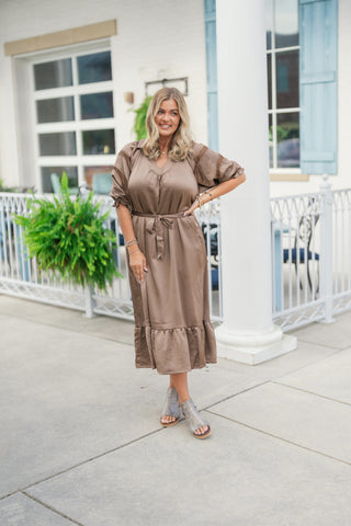 Woman posing outdoors smiling in a taupe mocha-toned satin midi dress, featuring an open V-neckline, raglan short sleeves with crochet lace inset panels, an adjustable self-tie waist sash, and a ruffled tiered hem. Styled with grey suede platform peep-toe sandals and delicate gold jewelry in front of a white building background.