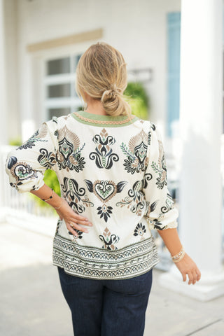 Back view of the off-white ikat printed blouse showing the round sage green trim along the back collar with tan scalloped lace detailing, relaxed three-quarter sleeves, and a straight border-print hemline. Styled over dark-wash jeans with hair styled in a loose low bun.