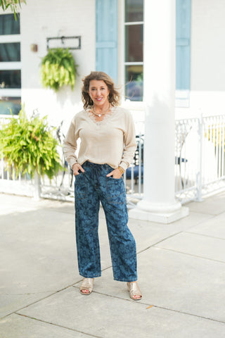 Woman posing outdoors wearing a light beige V-neck sweater featuring delicate eyelash lace trim along the neckline, paired with dark blue wide-leg denim jeans accented with an all-over tonal floral pattern. The casual chic outfit is styled with a drawstring waist, gold open-toe mules, a chunky gold chain necklace, and stacked gold bangles against a bright storefront backdrop.