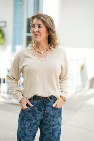 A portrait view highlighting the fine knit detail and intricate off-white lace trim frame of the light beige V-neck pullover sweater. The soft neutral sweater is tucked loosely into high-waisted floral print blue jeans featuring a relaxed drawstring tie and front patch pockets, accessorized with a gold pendant chain necklace and subtle drop earrings.