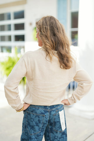Back view showcasing the smooth, continuous knit texture and long sleeves of the light beige sweater with hands resting on hips. Highlights the back fit of the blue floral print wide-leg jeans, displaying traditional back pockets and a comfortable relaxed silhouette.
