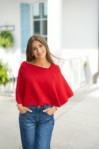 Woman smiling outdoors wearing a vibrant cherry red fine-knit poncho top featuring an asymmetrical slash neck, diagonal seam detailing across the front, and draped batwing-style cape sleeves. Styled with hands in the pockets of dark-wash wide-leg jeans and gold delicate jewelry.