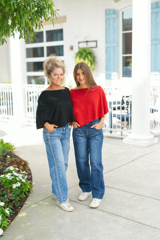 Two women posing side-by-side outdoors showcasing the fine-knit poncho top in both black and cherry red color options. The model on the left wears the black version paired with medium-wash straight-leg jeans, while the model on the right wears the red version paired with dark-wash wide-leg jeans, both styled casually with white sneakers.