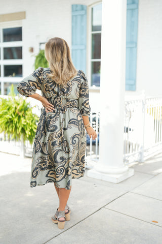 Back view of the sage green paisley printed midi dress highlighting the wide smocked elastic panel across the back waist for a comfortable, contoured fit. Features three-quarter puff sleeves, a fluid skirt silhouette hitting below the knee, and a subtle back yoke, styled with platform sandals.