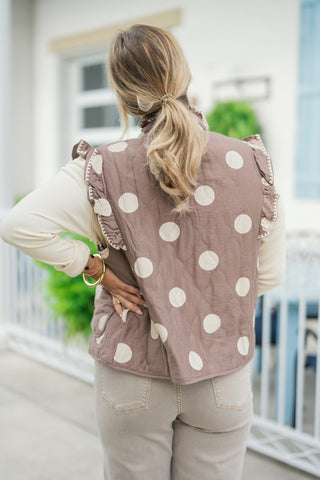 Back view of the taupe quilted vest showcasing the scattered cream polka dot pattern, ruffle trim detailing along the back shoulder seams, and a clean, straight hemline. Styled over a cream long-sleeve shirt and taupe jeans, viewed from behind with a low ponytail hairstyle.