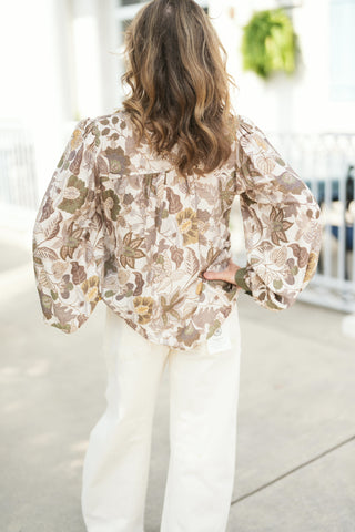 Back view of the tapestry floral print blouse showcasing the gathered back yoke for a comfortable, fluid drape and dramatic volume through the balloon sleeves. Styled with hands on hips, paired with high-waisted cream pants.