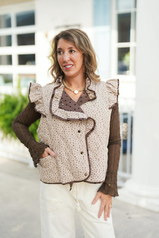 Woman smiling outdoors wearing a reversible quilted vest on the beige side, featuring a delicate brown ditsy floral print, ruffled shoulder wings, contrast piping, and a folded lapel revealing the reverse polka dot pattern. Layered over a sheer brown long-sleeve mesh top and cream wide-leg pants with a gold link necklace.