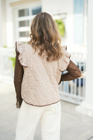 Back view of the reversible quilted vest shown on the beige ditsy floral side, highlighting the dramatic ruffled armhole trim edged with contrast piping. Layered over a brown textured long-sleeve shirt and paired with off-white denim pants.