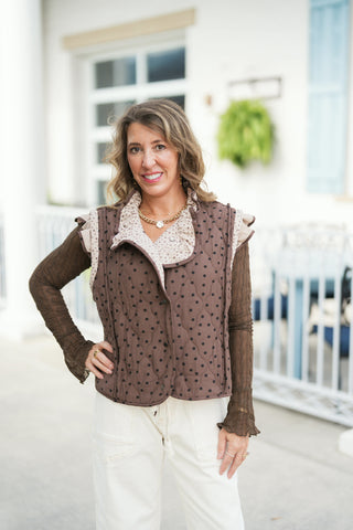 Front view of the vest reversed to show Side B, crafted in a chocolate brown quilted fabric with black polka dots. Features a snap-button front, ruffled trim along the armholes with the floral pattern showing, and a relaxed silhouette styled over a brown long-sleeve top and cream pants.