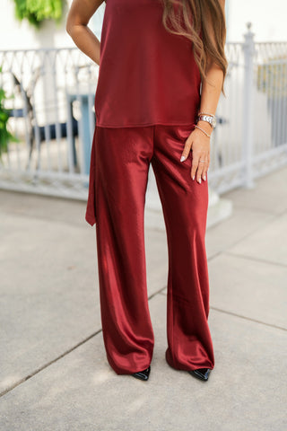 Lower-body shot of a woman modeling deep burgundy satin wide-leg pants with a glossy, high-shine finish. The trousers feature a high-waisted flat front and a relaxed, fluid drape through the leg, paired with matching burgundy satin halter top and black pointed-toe heels on an outdoor concrete walkway.