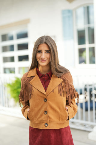 Woman smiling outdoors wearing a camel brown faux-suede Western jacket featured with classic notched lapels, dark brown front buttons, side welt pockets, and statement fringe trim cascading across the upper chest and shoulders. Layered over a rich burgundy button-down shirt and matching burgundy skirt for a chic, autumnal Western-inspired outfit.