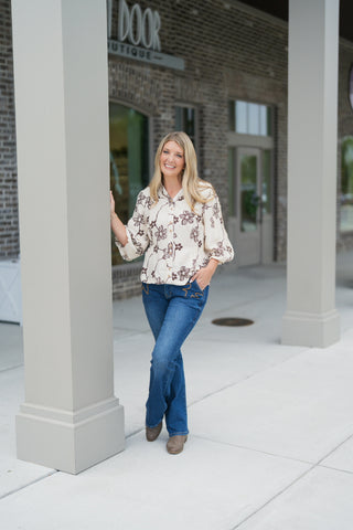 Woman standing outdoors in front of a modern brick storefront, posing with one hand leaning against a building pillar and the other in her pocket. She is wearing a cream-colored woven blouse featuring brown line-art floral embroidery, 3/4-length balloon sleeves, and ornate gold front buttons, paired with medium-wash bootcut jeans and tan suede ankle boots.