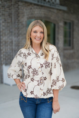 Upper-body portrait of a smiling woman posing with hands on her hips, wearing the cream and brown floral embroidered blouse. The image captures the split V-neckline collar, structured button-down front, wide elbow sleeves, and gold hoop earrings against a blurred storefront background.