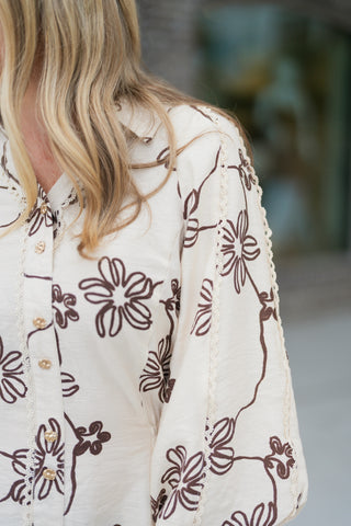 Close-up shot highlighting the fine craftsmanship of the cream blouse, showcasing the textured brown embroidered flowers, vertical crocheted lace trim along the button placket and shoulder seams, and vintage-inspired metallic gold crest buttons.
