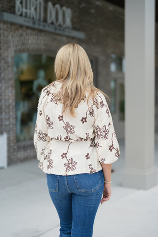 Back view of the cream embroidered blouse showing the relaxed fit, smocked cinched waistline, and back floral embroidery pattern. The blouse features elbow-length voluminous sleeves with delicate crochet lace inset trim along the raglan shoulder seams, worn over medium-wash blue denim.