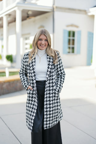A waist-up shot highlighting the structured notch lapels and tailored open-front silhouette of the black and white oversized houndstooth coat. Layered over a cream fine-ribbed turtleneck sweater accessorized with a delicate gold drop pendant necklace.