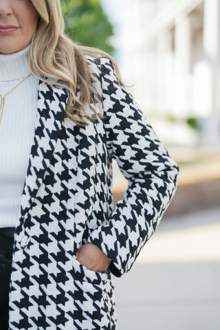Extreme close-up shot focusing on the tactile woven texture of the black and white houndstooth pattern fabric and the functional front side pocket. Shows the layering detail with a white ribbed turtleneck and gold necklace.