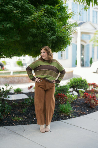 Woman posing in a olive green and chocolate brown color-blocked striped sweater, featuring relaxed drop-shoulder sleeves and textured eyelash yarn trim outlining each horizontal stripe. The slouchy knit is front-tucked into high-waisted caramel brown velvety wide-leg pants paired with two-tone beige ankle booties. Styled outdoors in front of a leafy green tree and modern building.   