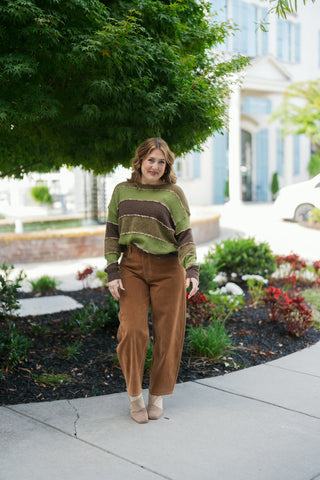 Full-length front view of the olive green and brown striped crewneck knit sweater with hands resting on the hips. Highlights the relaxed barrel-leg silhouette and exposed contrast stitching of the caramel-colored wide-leg trousers.