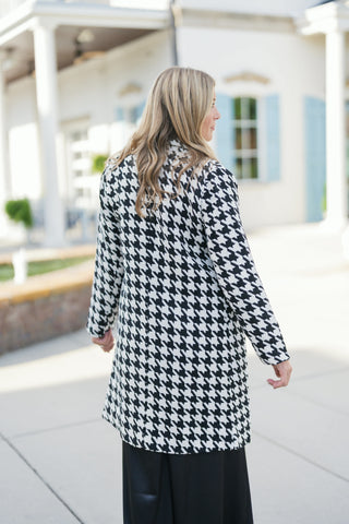 Back view showcasing the continuous black and white houndstooth pattern across the shoulders, back seam, and sleeves of the mid-length coat. Highlights the straight-line silhouette and hem coverage over black wide-leg pants.