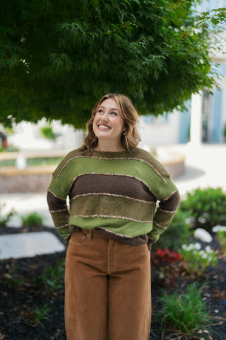 Portrait shot showcasing the crew neckline and subtle multi-color flecks in the fuzzy eyelash seam piping along the olive and brown knit stripes. The relaxed pullover top is styled with simple gold hoop earrings.