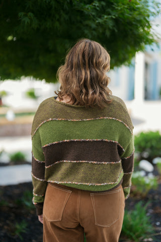 Back view of the striped knit sweater, demonstrating the seamless horizontal color-blocking in olive green and dark brown across the shoulders and lower back. Highlights the back patch pocket placement and white contrast stitching on the caramel brown wide-leg pants.
