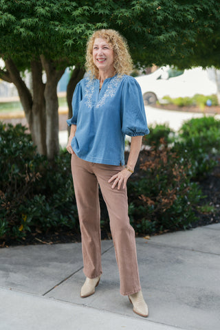 Woman posing outdoors in a light wash blue chambray blouse featuring delicate white floral embroidery around a split neck slit collar and voluminous elbow-length puff sleeves with elastic cuffs. The relaxed top is paired with warm taupe straight-leg pants and pointed-toe snakeskin ankle boots. Styled with blonde curly hair in front of a lush green tree.