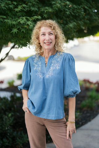 A three-quarter front view highlighting the bohemian white floral embroidery framing the V-notch neckline and gathered sleeve shoulders of the light denim-look chambray top. Styled untucked with one hand resting in the pocket of dusty tan pants.