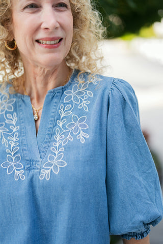 A detailed portrait shot emphasizing the intricate white thread floral and vine embroidery surrounding the split V-neck placket. Accessorized with a delicate gold pendant necklace and gold hoop earrings.   