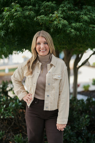 A portrait view showcasing the detail of the cream corduroy jacket with silver snap buttons, chest flap pockets, and distressed raw-edge shoulder seams. Layered over a fitted taupe ribbed turtleneck sweater with a gold necklace and paired with high-waisted dark brown pants.
