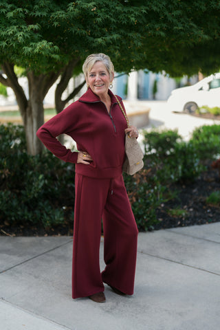 Woman posing outdoors in a rich burgundy monochrome lounge set featuring a long-sleeve half-zip pullover sweatshirt with an exaggerated spread collar and matching high-waisted wide-leg sweatpants. Styled with a taupe quilted suede shoulder bag with a gold chain strap, gold statement rings, and brown suede flats under a lush green tree backdrop.