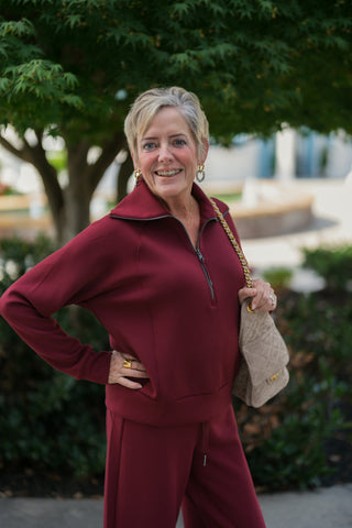 A portrait view highlighting the smooth, structured knit texture of the wine-red half-zip pullover sweatshirt. Features an open silver zipper front, dramatic folded collar, raglan sleeve seams, and a ribbed waistband, styled with a taupe quilted suede purse and gold drop earrings.