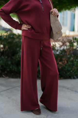A detailed view from the bust down highlighting the smooth, structured knit texture of the wine-red half-zip pullover and wide-leg trousers. Shows the front silver zipper detail, ribbed waist hemband, elastic drawstring waistband with metal aglets, and the taupe quilted suede purse tucked under the shoulder.